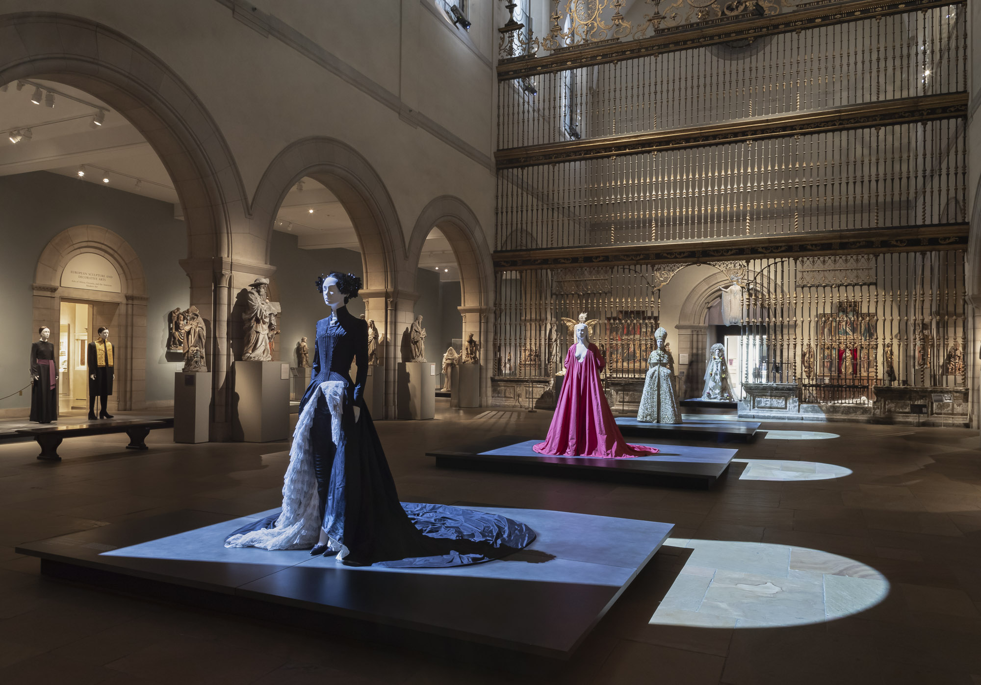 A procession of mannequins in long couture gowns stand atop four large, low, steel platforms lining the center of Medieval Art Galleries at The Met Fifth Avenue.